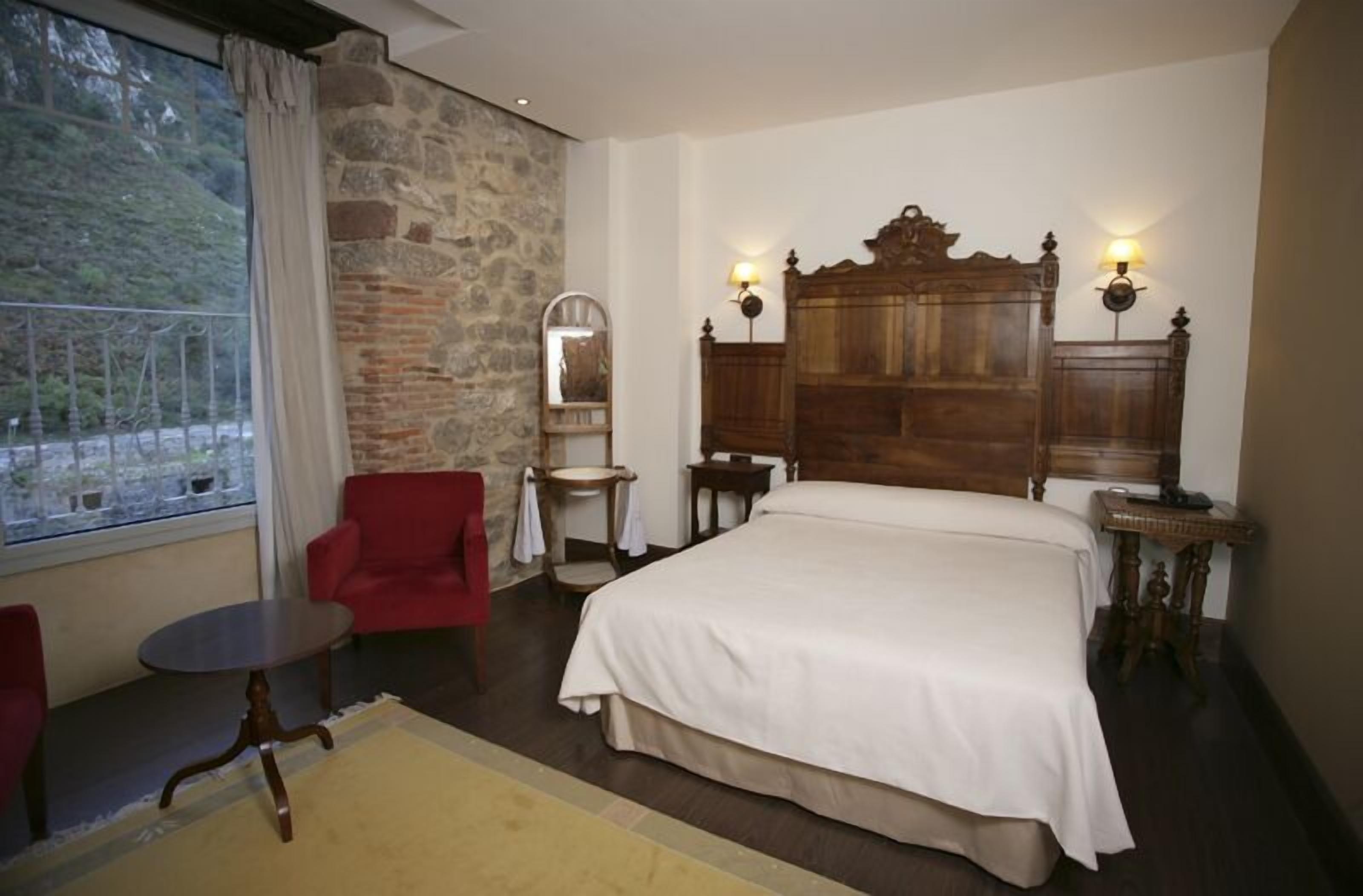 A hotel room with a large bed, a red armchair, a small round table, and a stone wall.