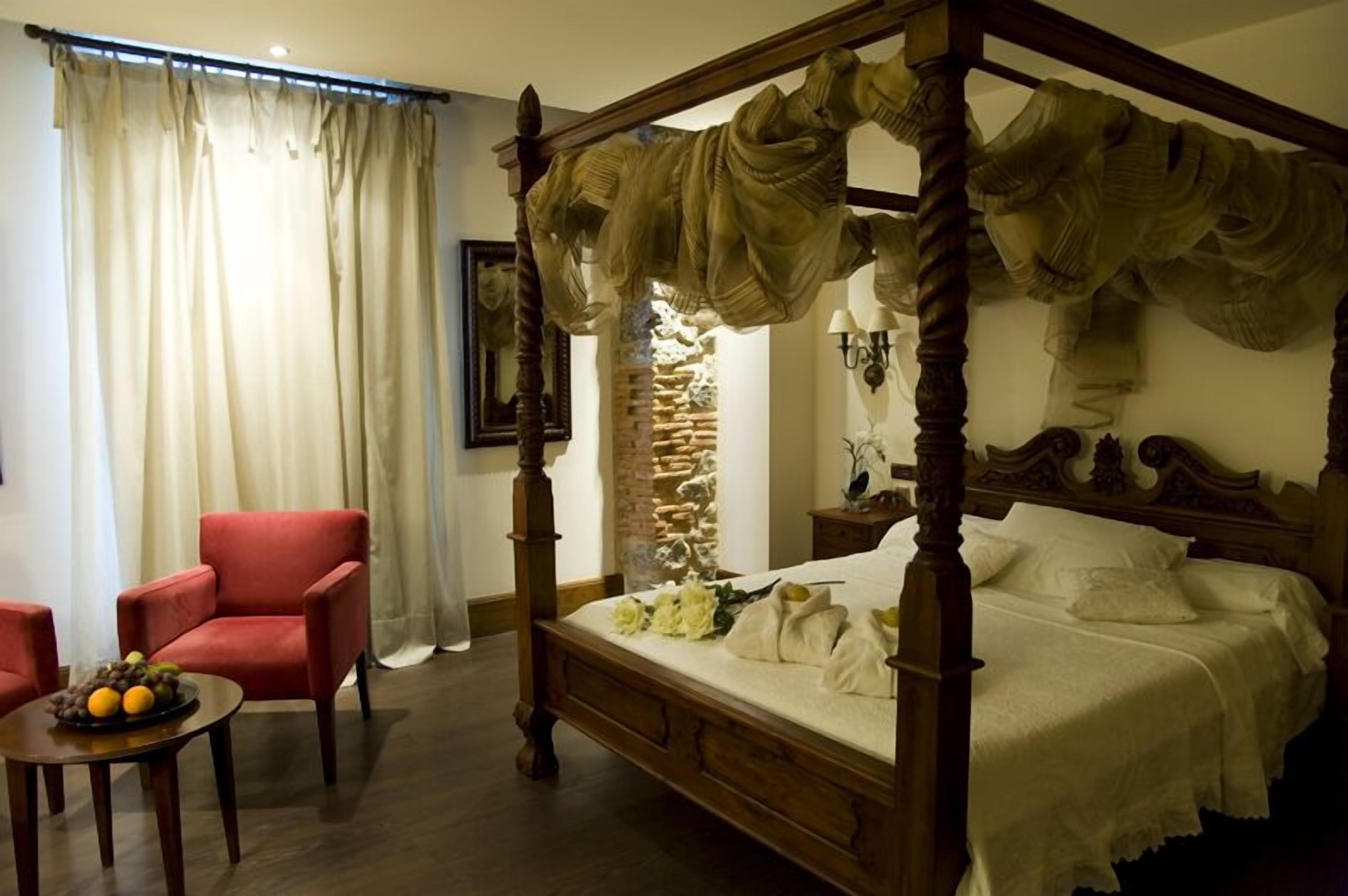 Una cama con dosel frente a una pared de piedra, un sillón rojo y una mesita con fruta.