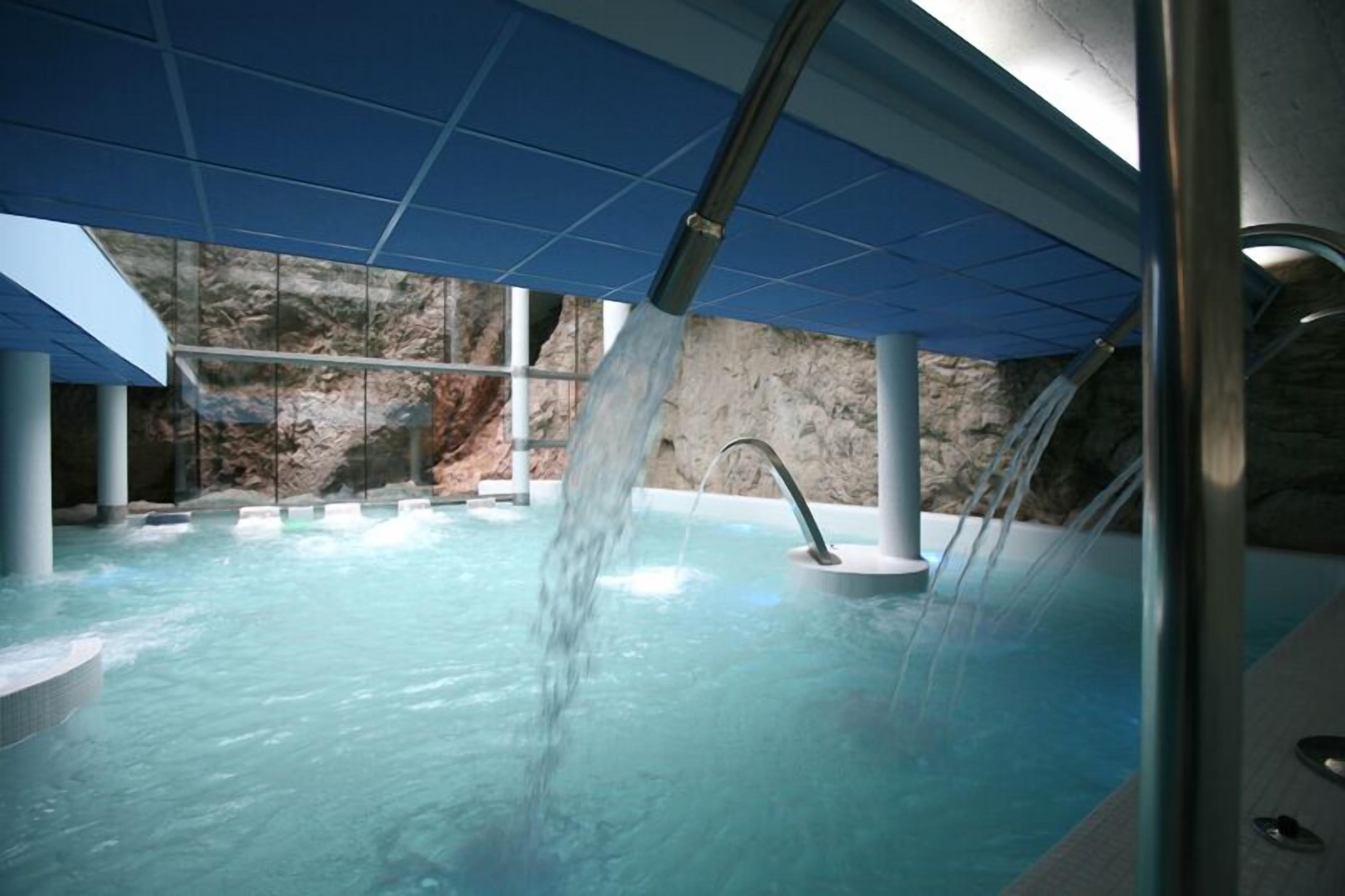 A modern indoor pool with clear water, a waterfall feature, and a view of a rocky landscape through large windows.