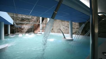 A modern indoor pool with clear water, a waterfall feature, and a view of a rocky landscape through large windows.