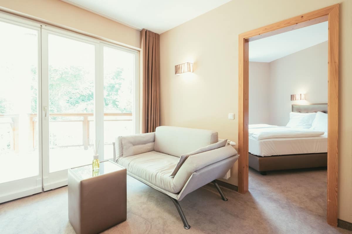 A modern hotel room with a large sliding glass door leading to a balcony, a comfortable sofa, a small table, and a neatly made bed.