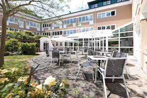 An outdoor patio area with tables and chairs, surrounded by a building with large windows and a stone pathway. - Hotel TraumVogel (Jennersdorf)