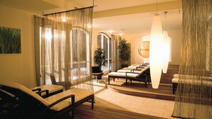 Un moderno salón de spa con suelo de madera, una zona de estar con sillas y un sofá, un espejo grande y una lámpara colgante.