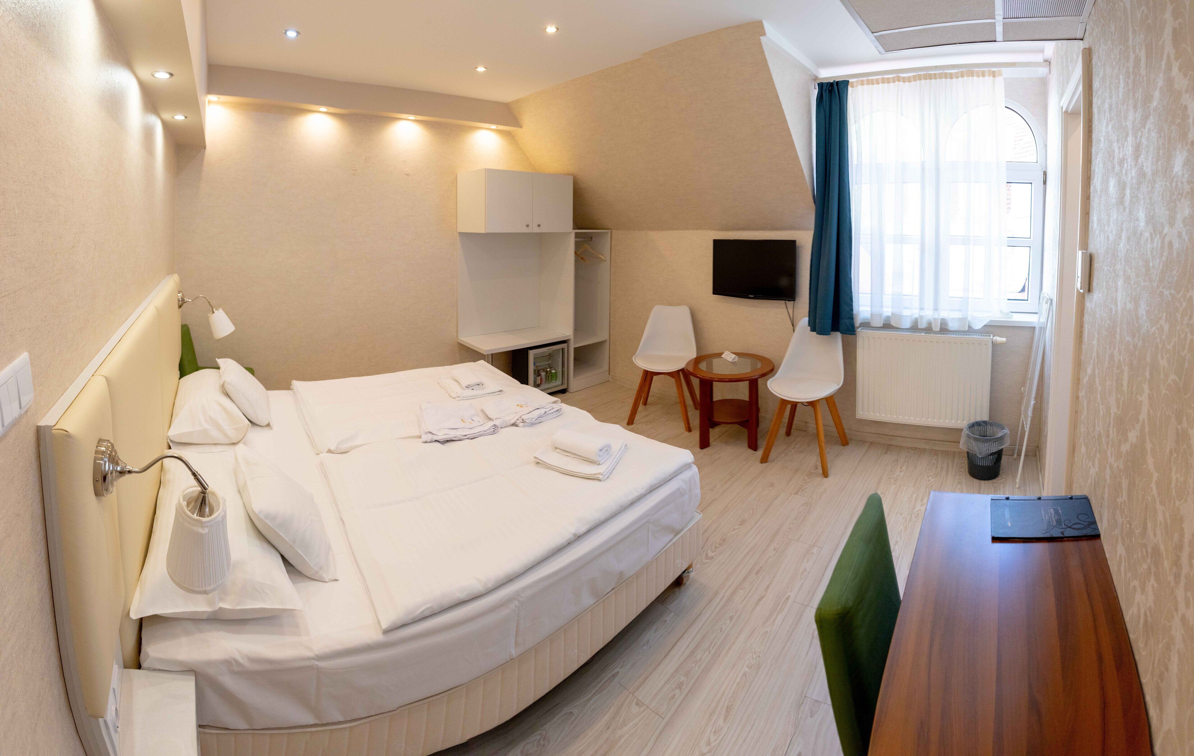 Habitación de hotel con cama, mesita de noche, silla, televisor y una pequeña mesa con otra silla.