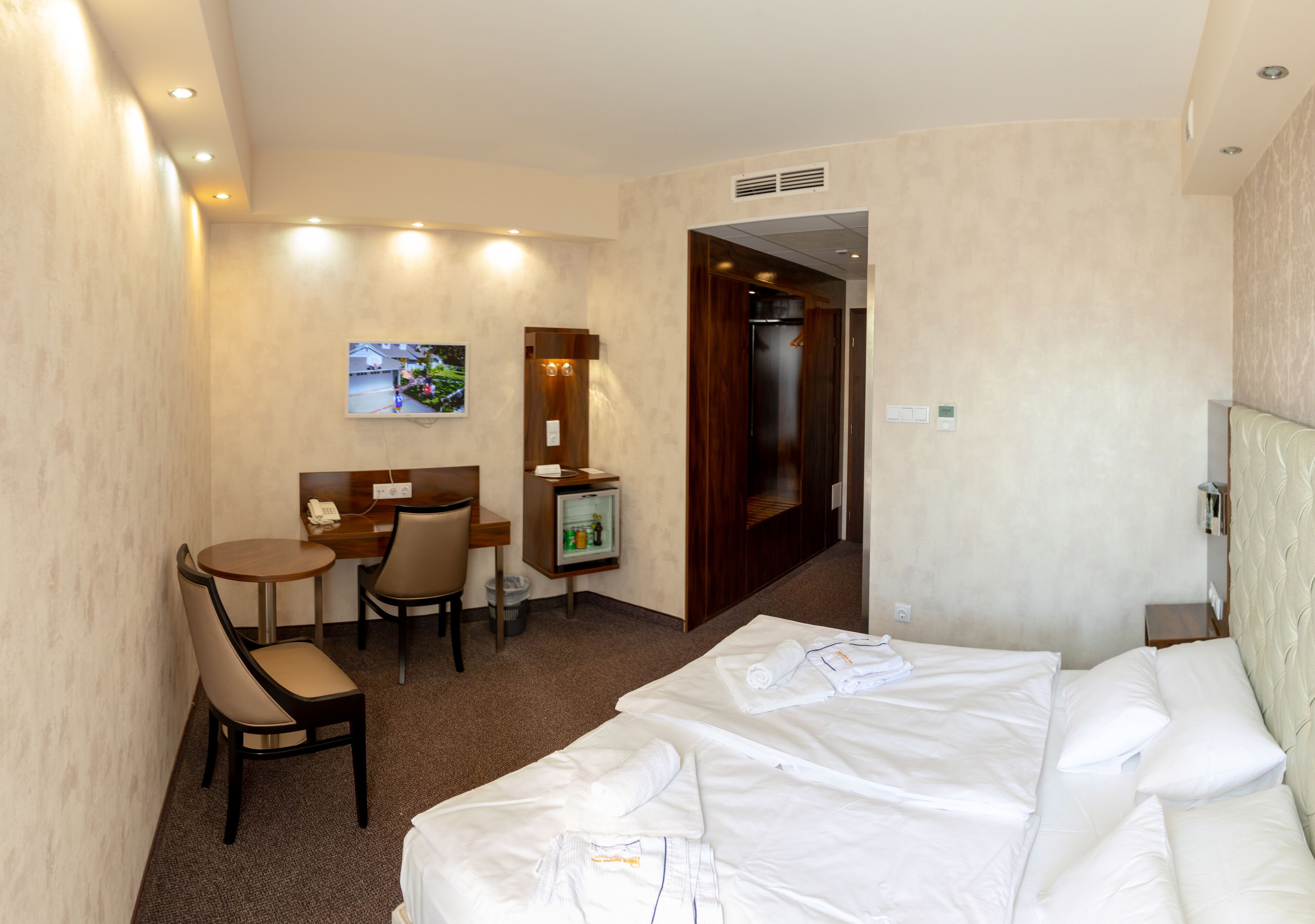 A hotel room with a bed, a desk with a chair, a small refrigerator, and a television on the wall.