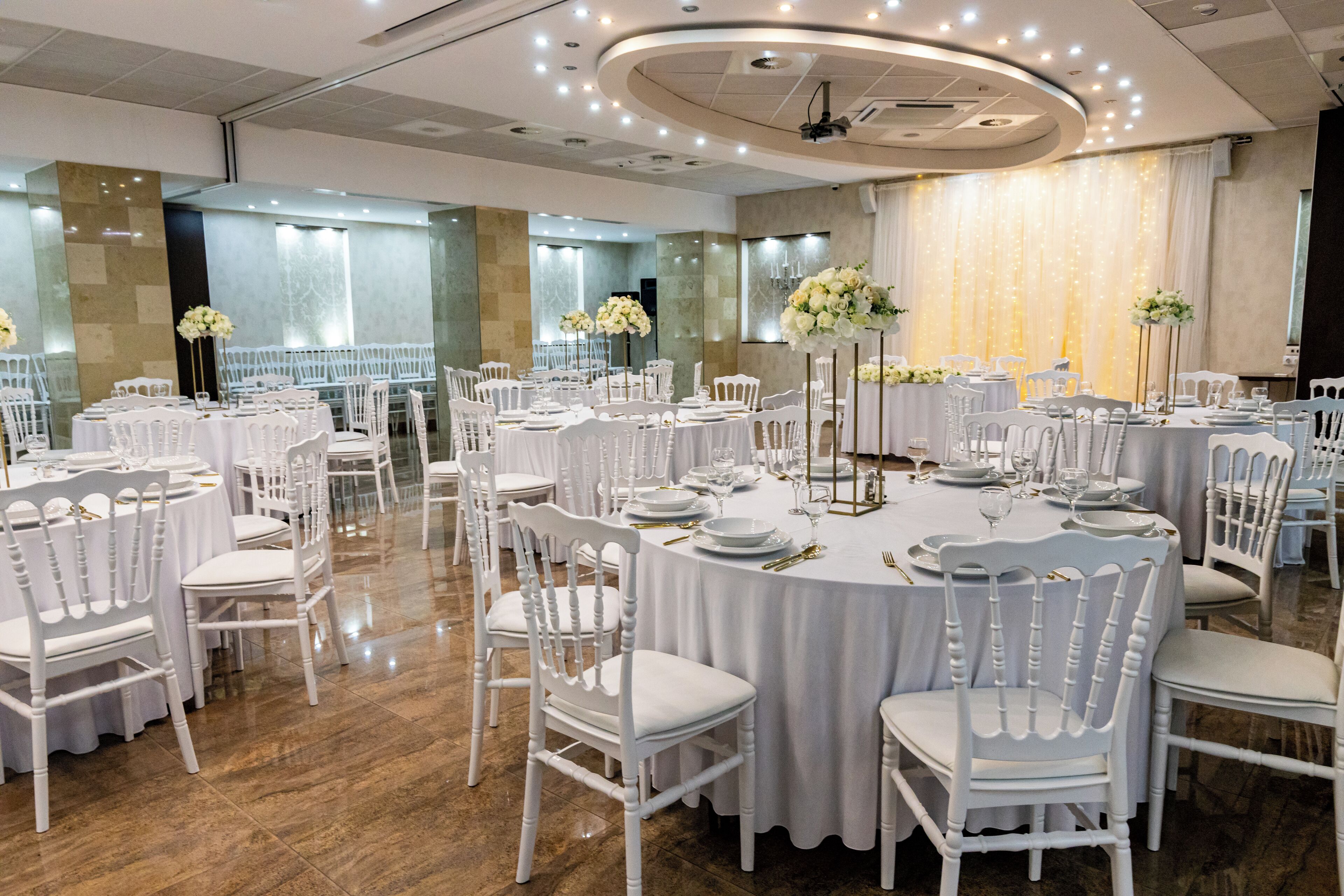 A banquet hall with round tables, white chairs, and floral centerpieces.
