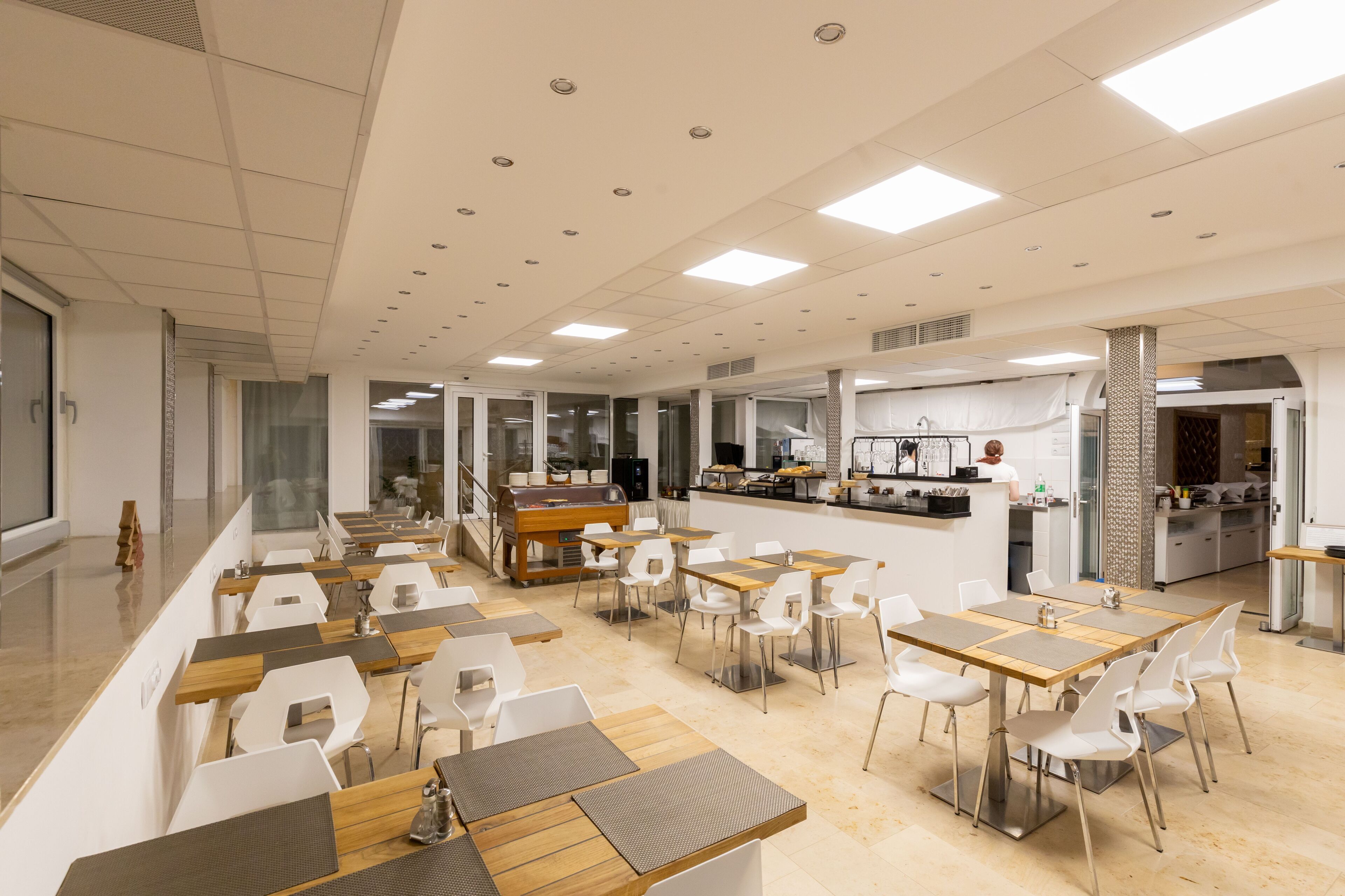 Un comedor amplio con varias mesas y sillas, una cocina al fondo y vistas de la ciudad a través de amplias ventanas.
