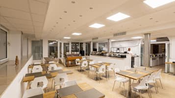 Un comedor amplio con varias mesas y sillas, una cocina al fondo y vistas de la ciudad a través de amplias ventanas.