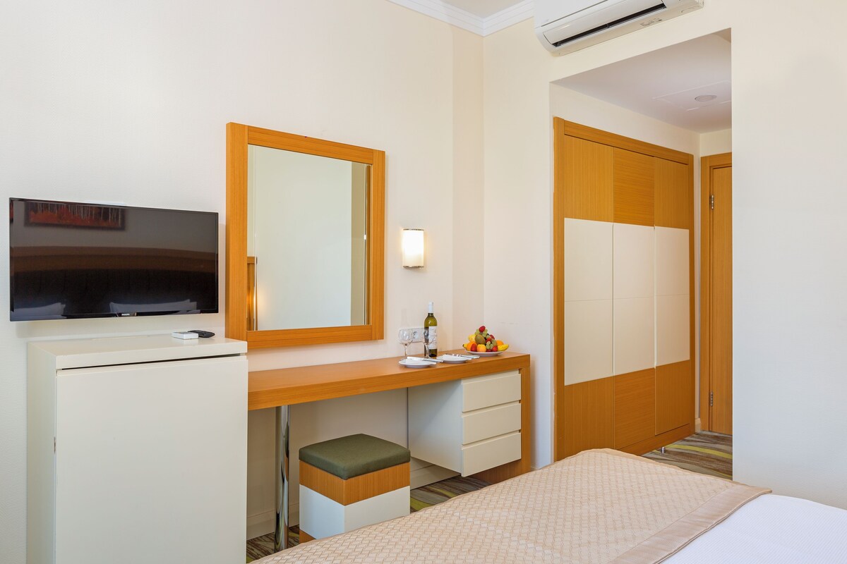 Une chambre d’hôtel comprenant un lit, un bureau avec une télévision, un miroir, un petit tabouret, une table de chevet avec une bouteille et des verres, et une armoire.