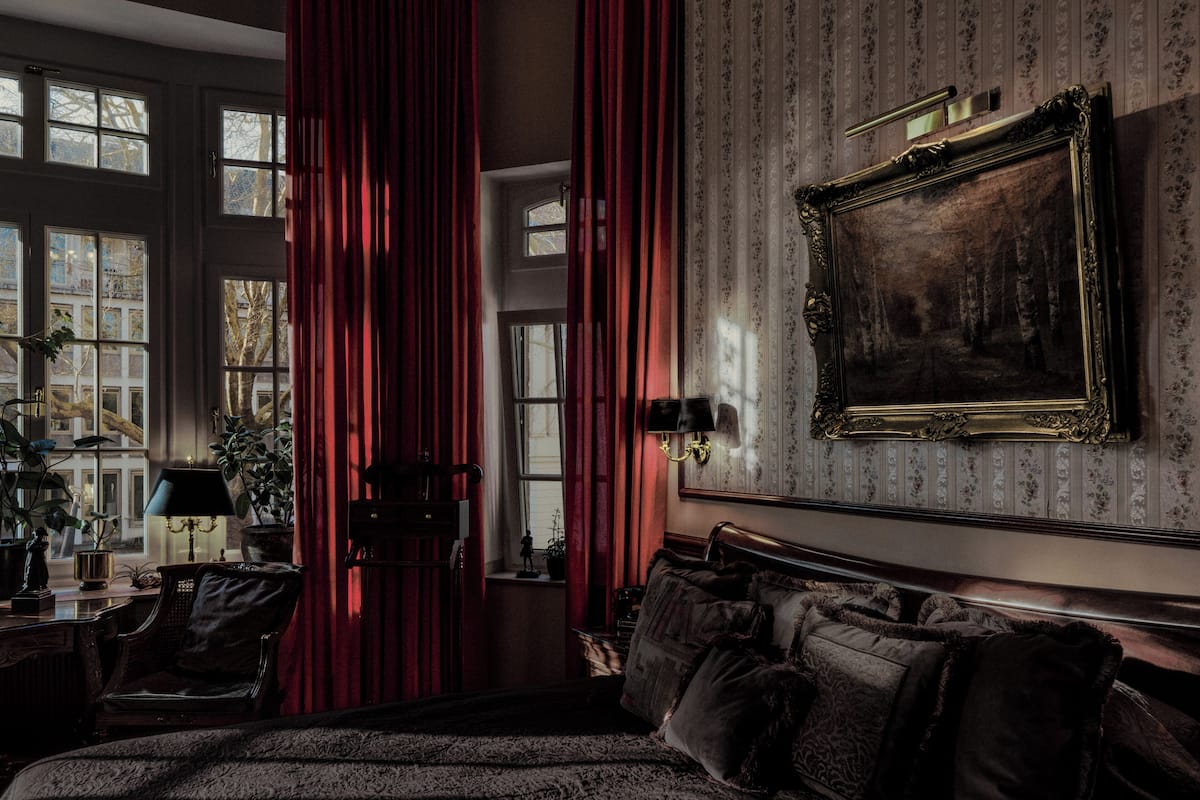 A room with a large bed, a desk with a lamp, a chair, and a large window with curtains.