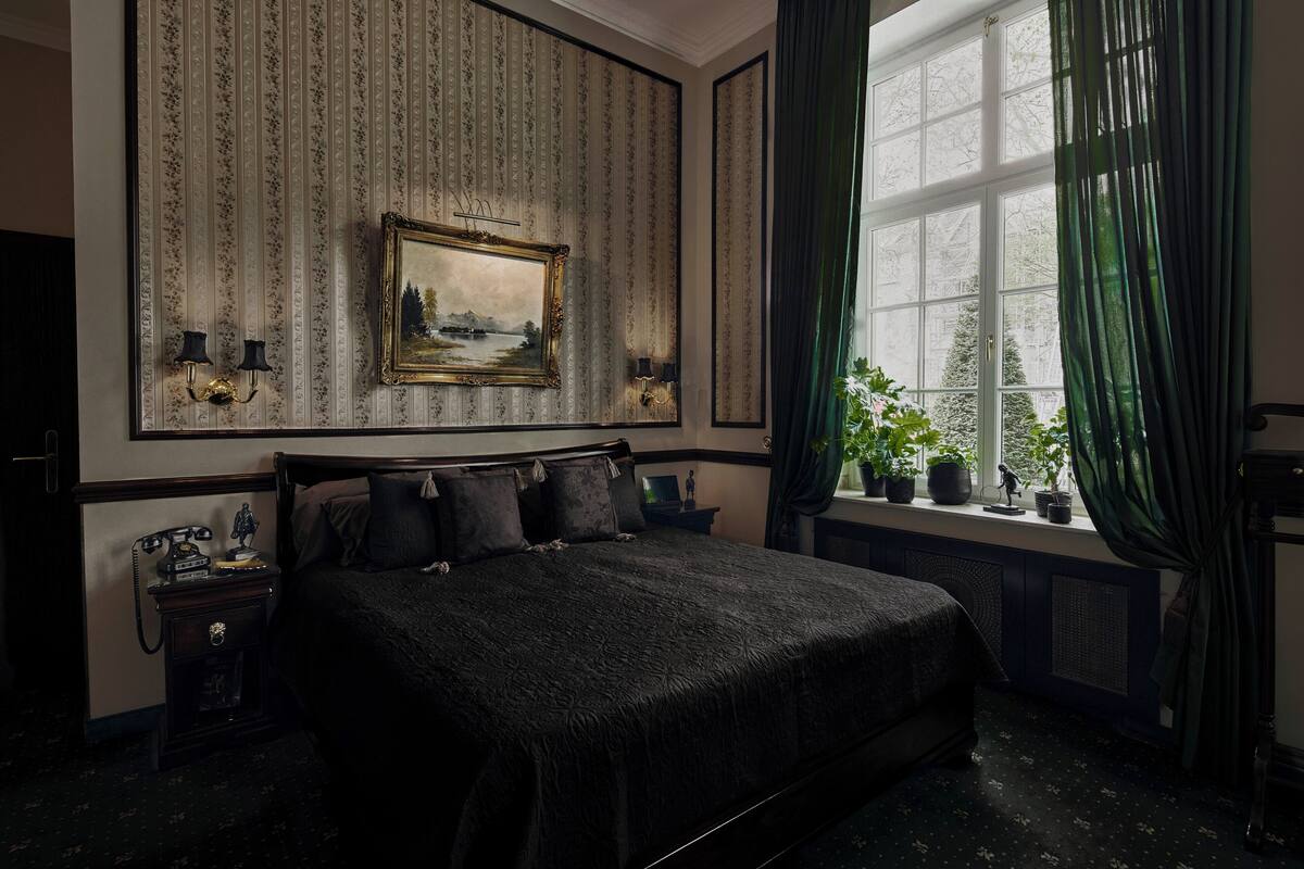 A classic hotel room with a large bed, a nightstand with a telephone, a framed painting, and a window with curtains.