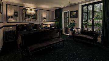 A dark-themed room with a large bed, a bench, a sofa, and a table with lamps. There is a large window with curtains and a view of greenery outside.