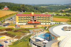 A multi-story hotel with a swimming pool, outdoor seating, and a view of the surrounding countryside. - Thermenhotel PuchasPLUS (Stegersbach)