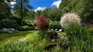 Un jardín exuberante con un estanque, rodeado de diversas plantas y árboles. Varias sillas de descanso están dispuestas para relajarse.
