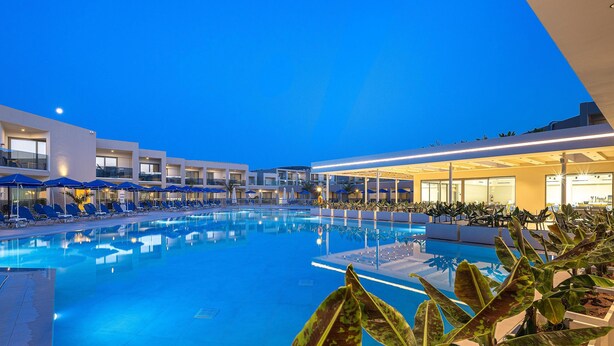 Una piscina con sillas de descanso y sombrillas, rodeada de edificios con grandes ventanales y un cielo nocturno.