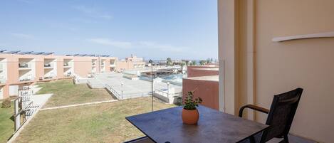 A balcony with a table and chairs, a potted plant, and a view of buildings and a grassy area.