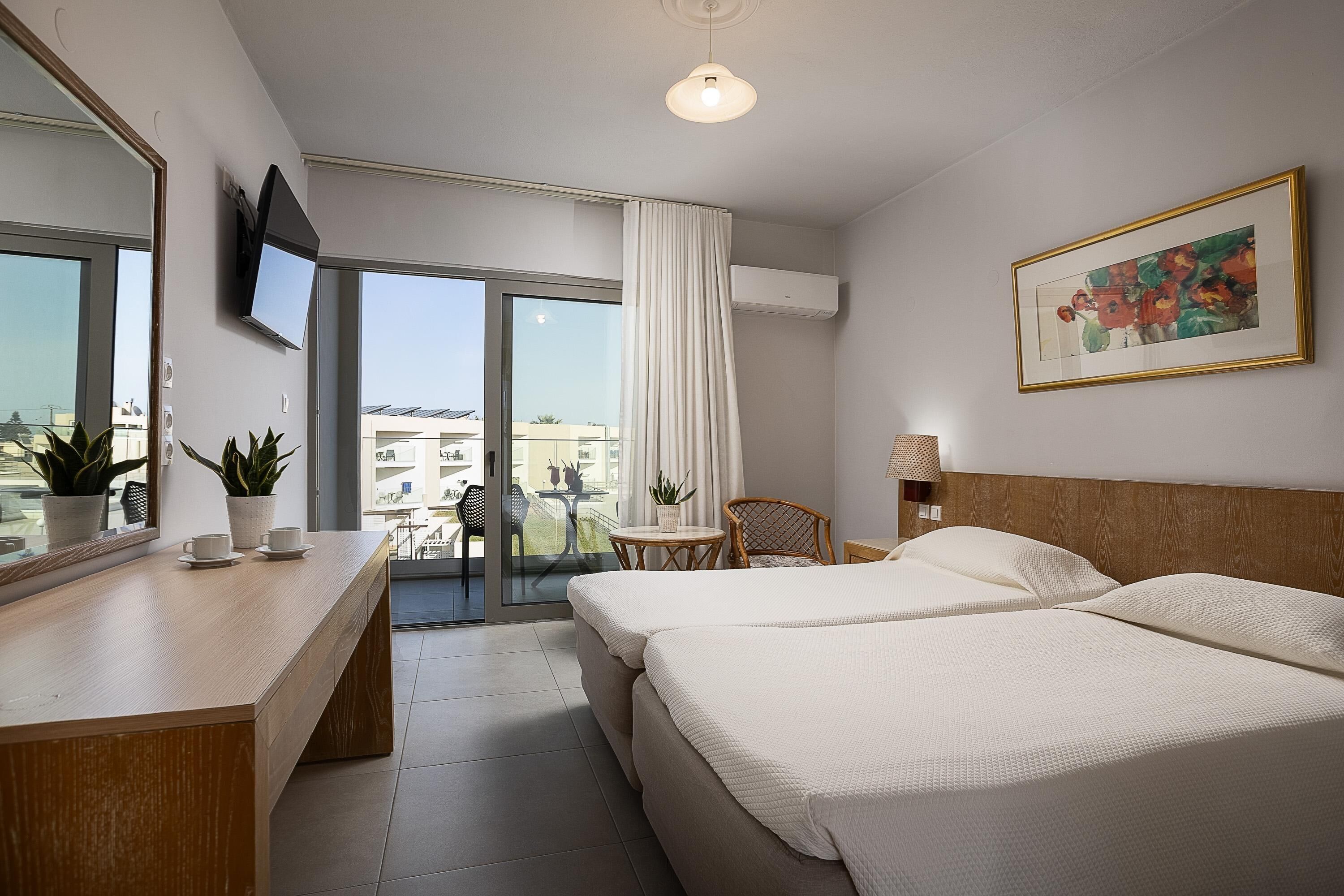 A hotel room with two beds, a desk with a mirror, a TV, a painting, and a balcony with a view of buildings.