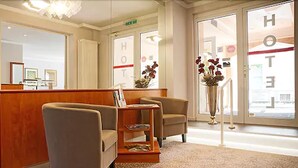 A hotel reception area with a wooden reception desk, two armchairs, a small coffee table, and a large window with a view of the street. - Hotel Victoria (Bad Kreuznach)