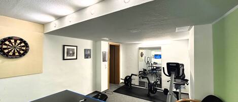 Una habitación con dardos, cuadros en la pared, mesa de ping pong, equipo de ejercicio y una televisión al fondo.