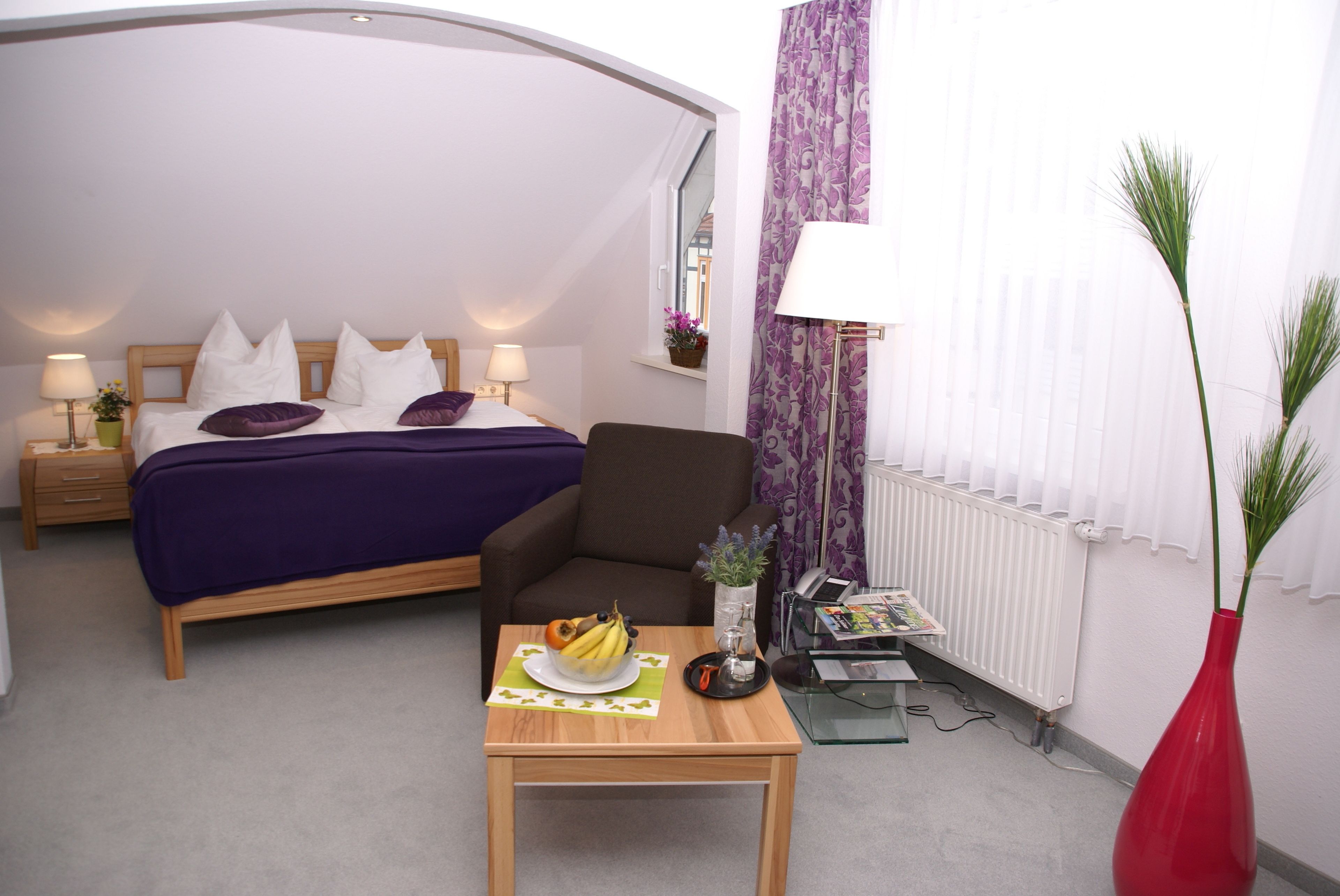 A bedroom with a bed, a chair, a table with fruit, a vase, and a window with curtains.