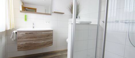 Un baño moderno con tocador de madera, cabina de ducha de vidrio y suelo de baldosas blancas.