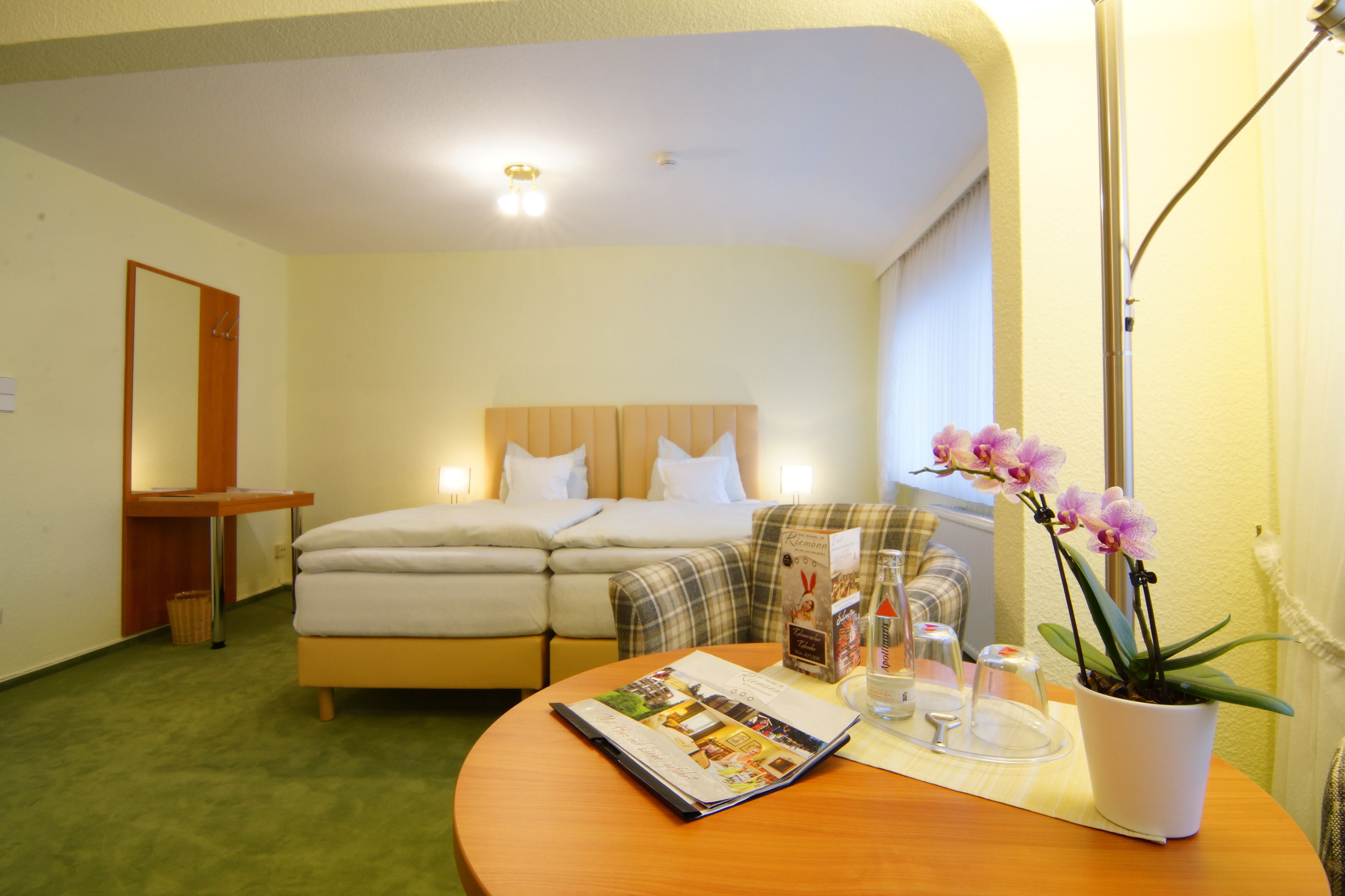 A hotel room with a bed, a bedside table with a lamp, a small table with a magazine and a potted orchid, and a window with curtains.