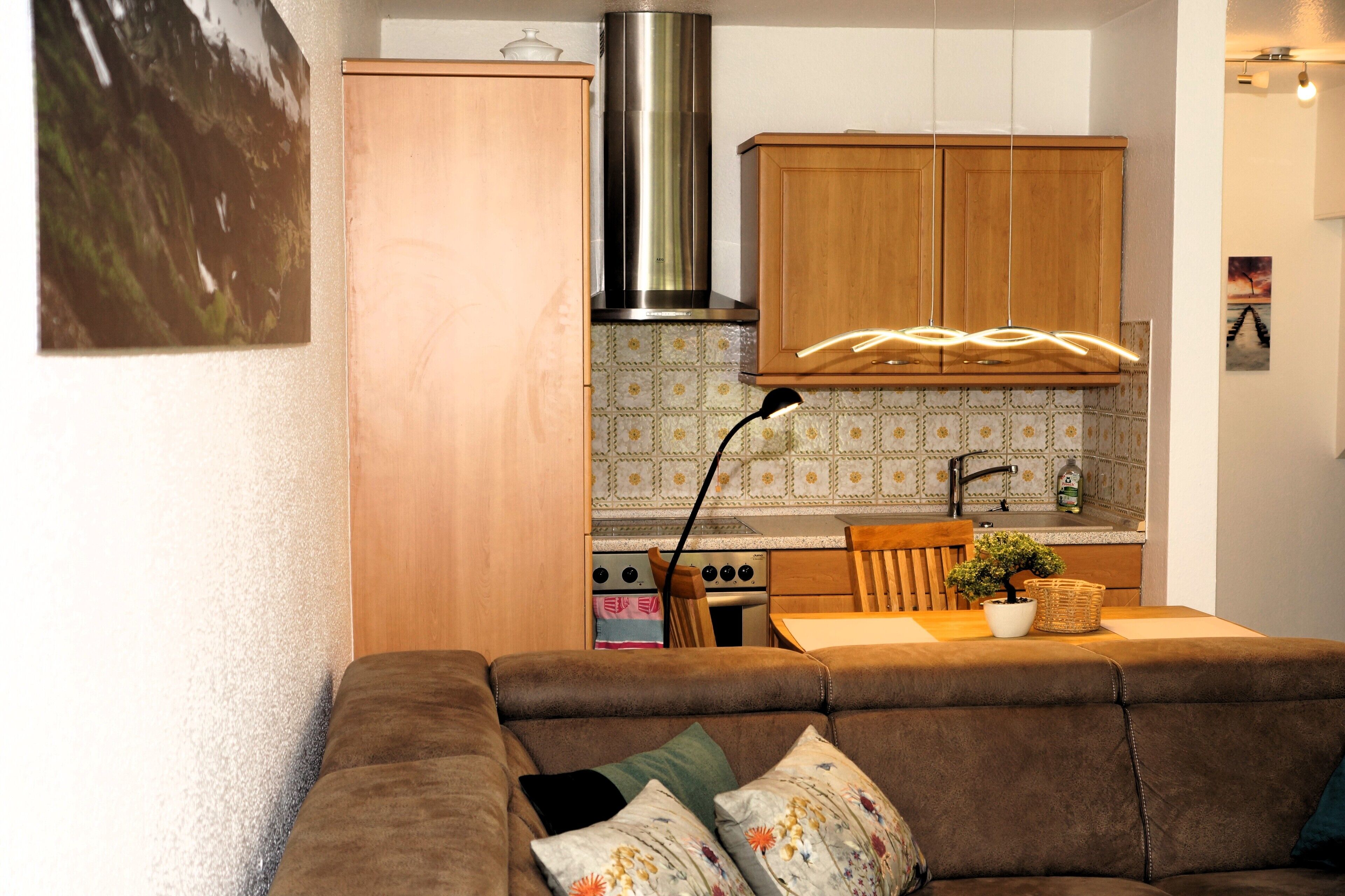A compact living space with a sofa, wooden cabinets, a kitchen area with a stove and sink, and a hanging light fixture.