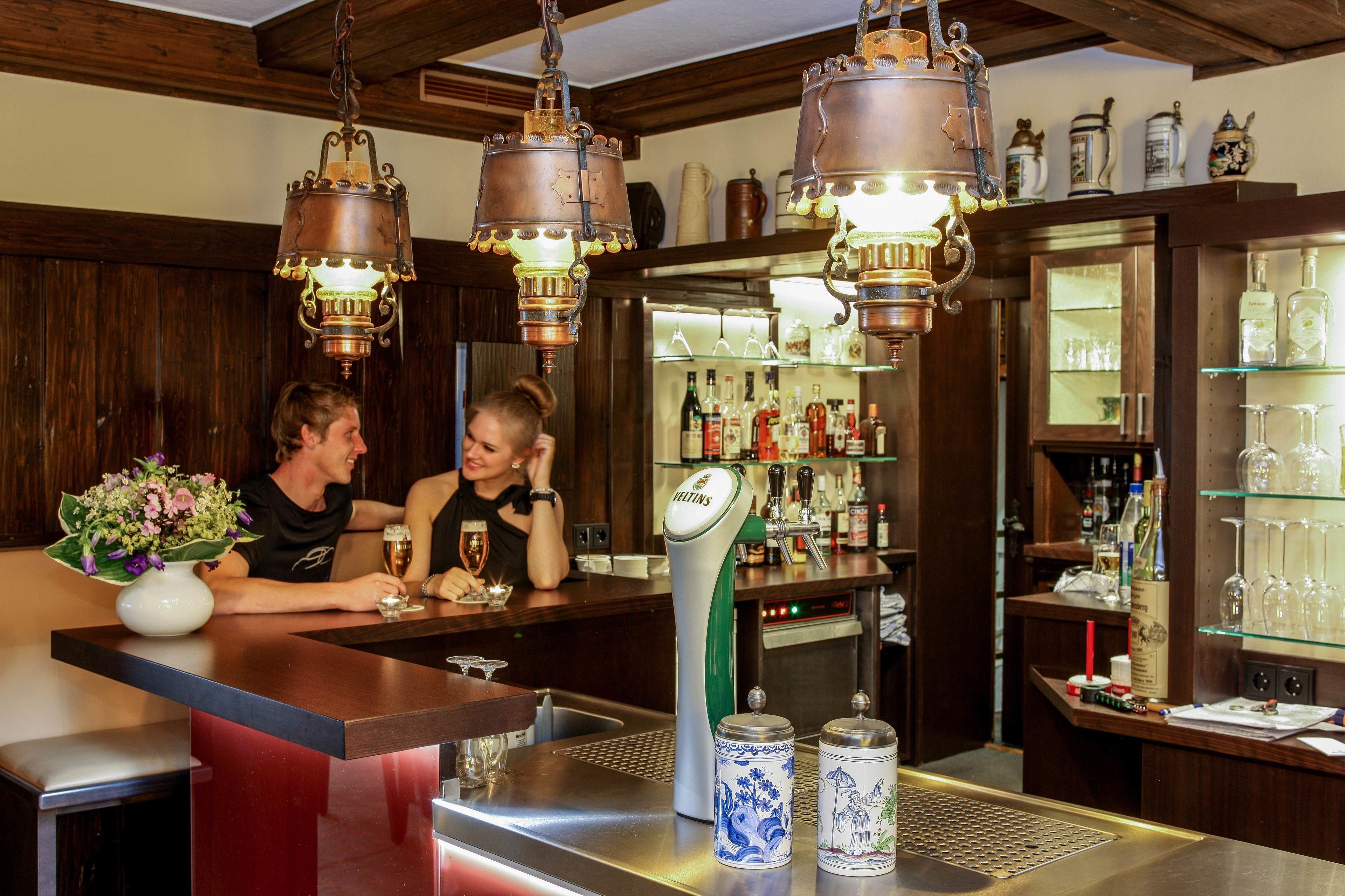 Un espace bar avec des lambris en bois, des lustres en cuivre suspendus et une étagère bien garnie de bouteilles et de verres.