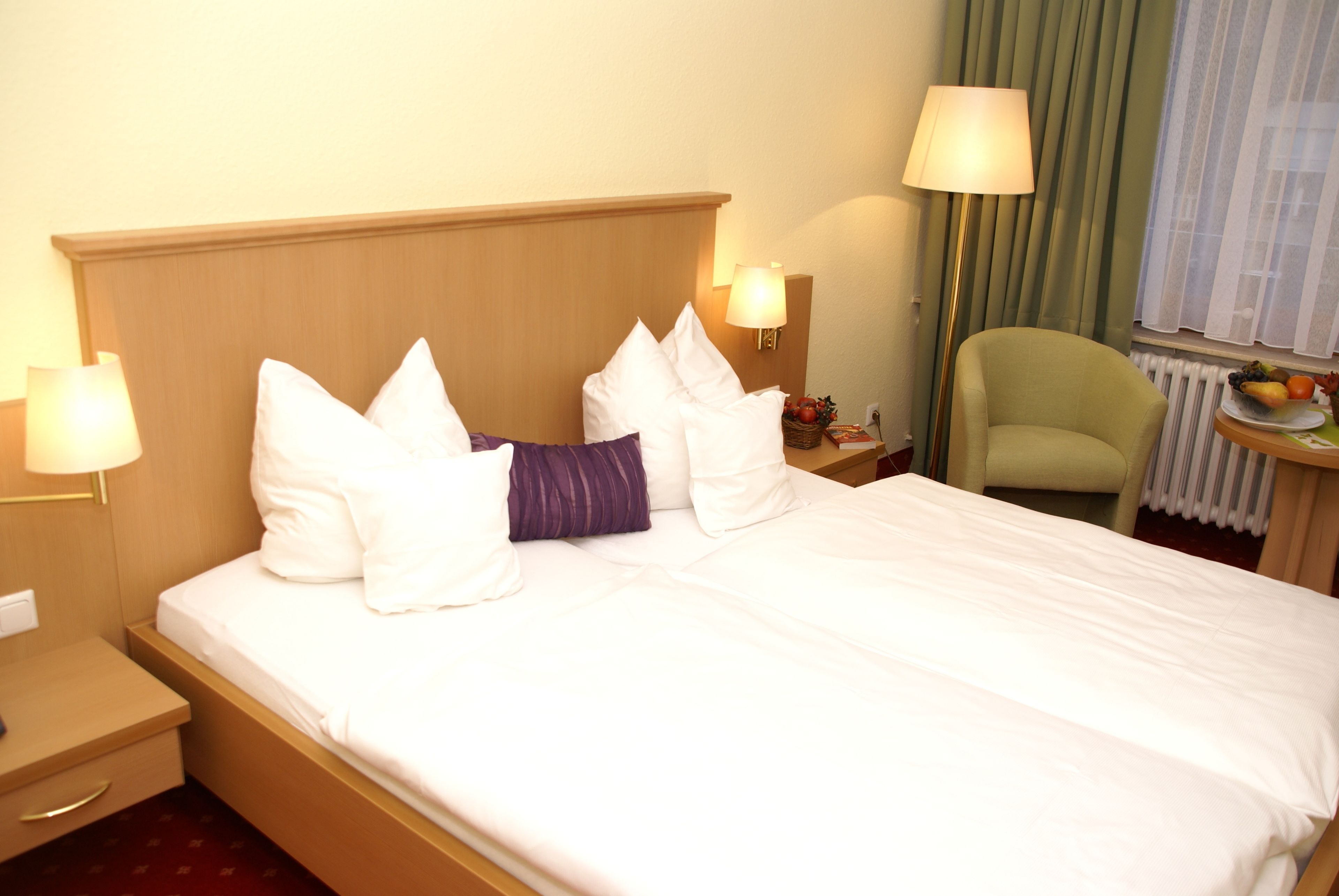 A hotel room with a neatly made bed, two bedside lamps, a chair, and a small table with fruit.