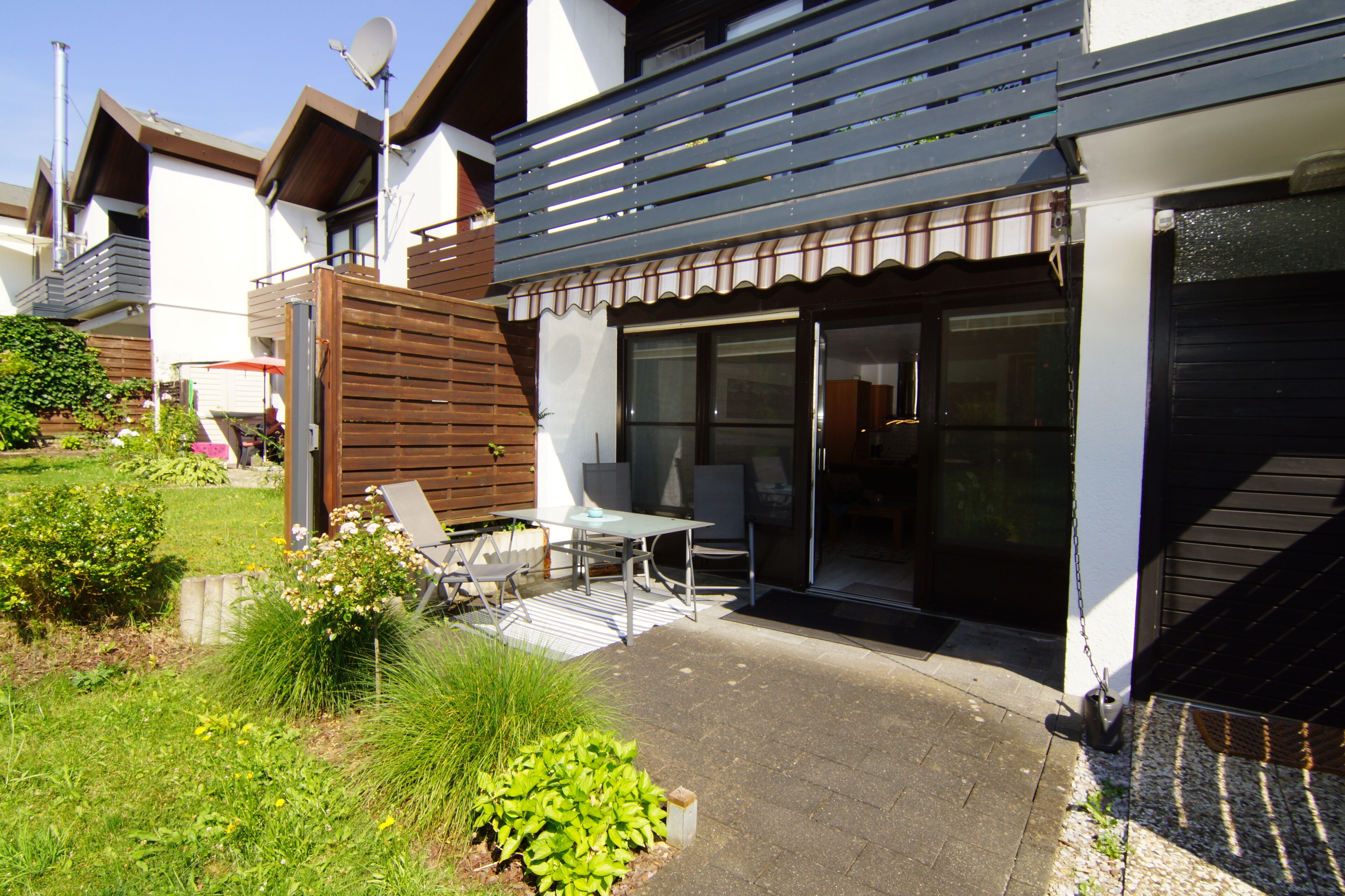 A modern house with a patio area featuring a table and chairs, surrounded by greenery and a wooden fence.