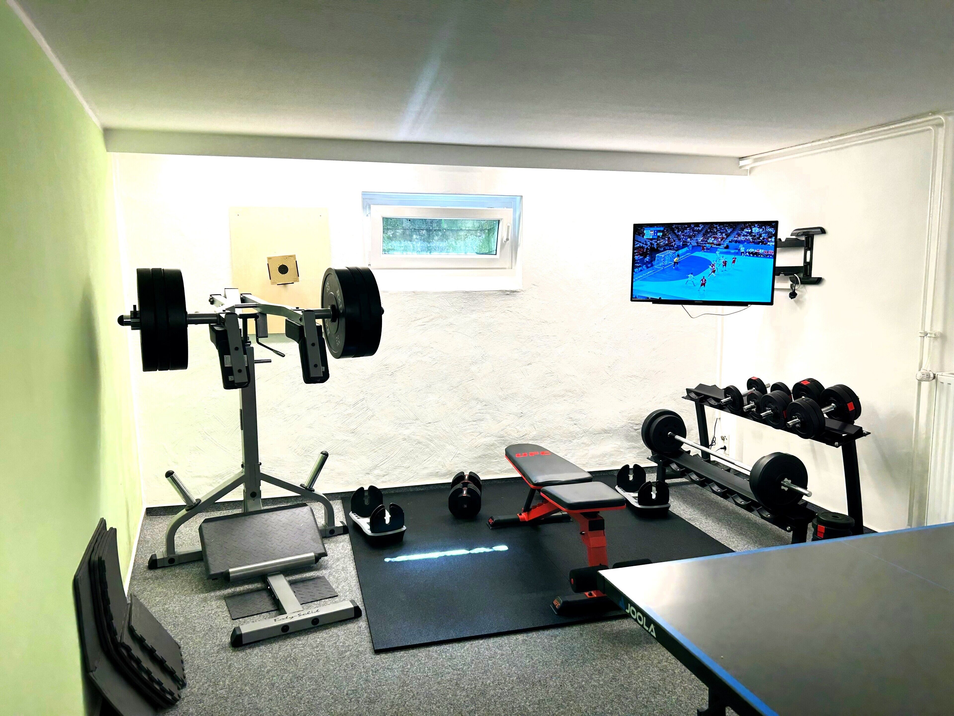 A home gym with a bench press, squat rack, dumbbells, and a TV displaying a sports event.