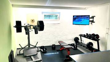 A home gym with a bench press, squat rack, dumbbells, and a TV displaying a sports event.