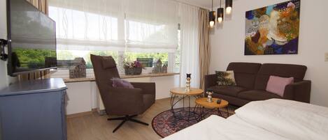 A modern hotel room with a bed, a sofa, a TV, a small table, and a window with curtains.
