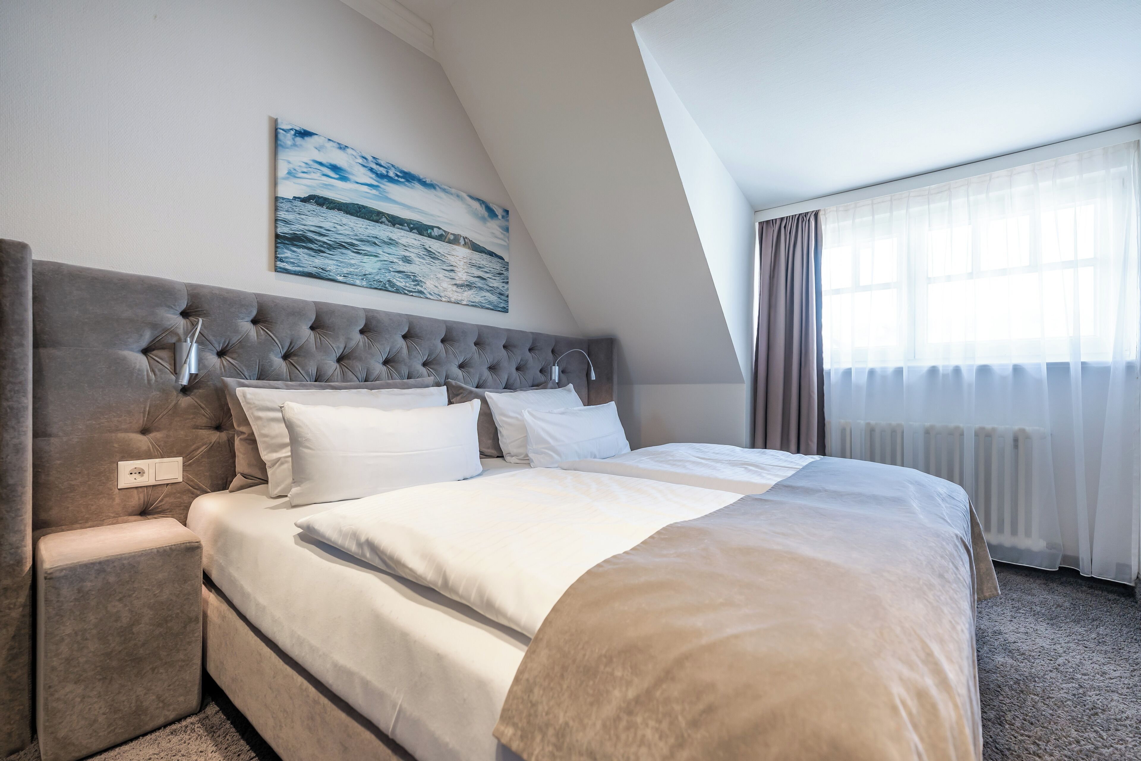 A hotel room with a large bed, a tufted headboard, a nightstand, and a painting of a seascape.