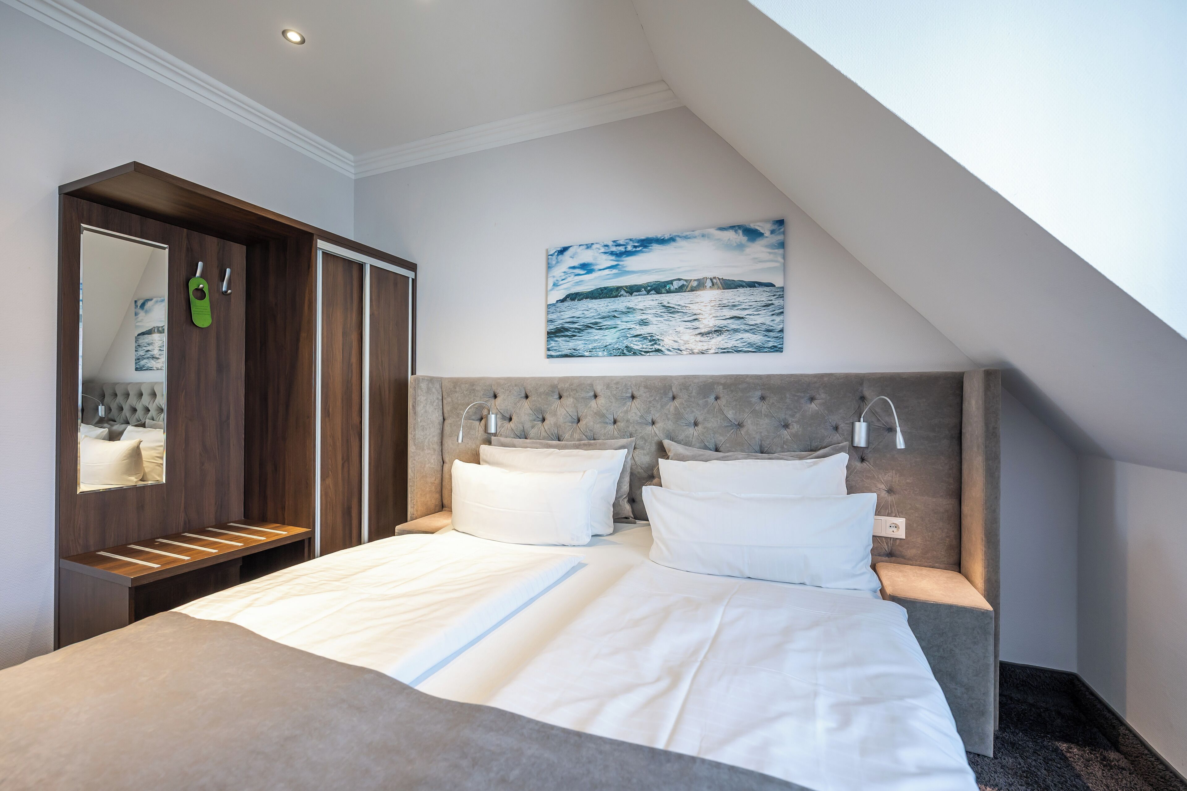 A modern hotel room with a large bed, a built-in wardrobe, a painting of a seascape, and a skylight.