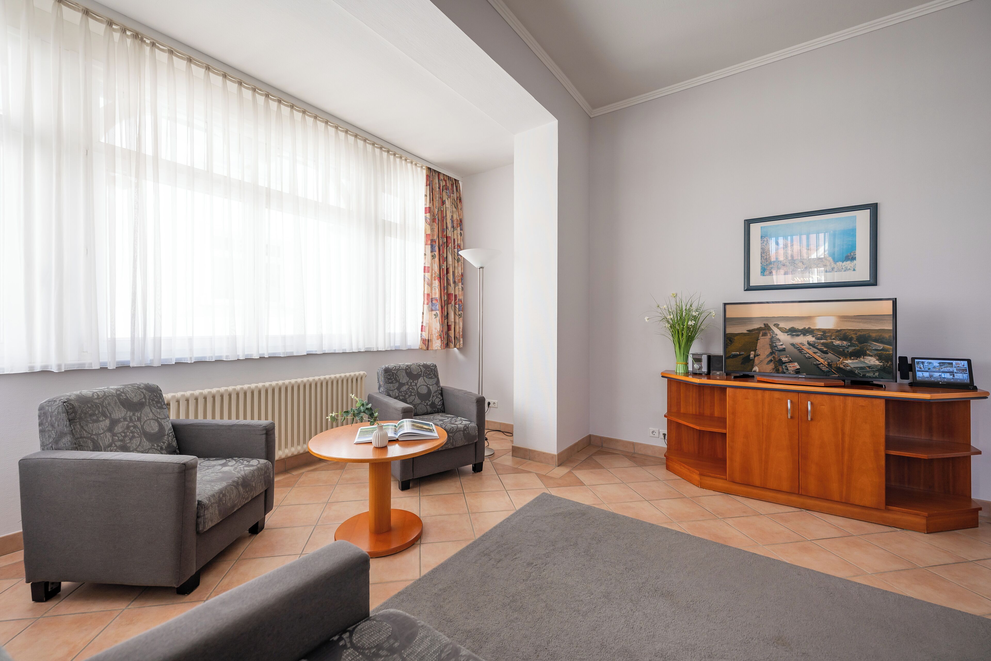 Una sala moderna con dos sillones grises, una pequeña mesa redonda con un libro, un mueble de madera con un televisor de pantalla plana y un ventanal con cortinas translúcidas.