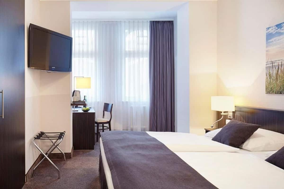 A hotel room with a bed, a desk with a chair, a TV mounted on the wall, a window with curtains, and a painting on the wall.