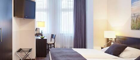 A hotel room with a bed, a desk with a chair, a TV mounted on the wall, a window with curtains, and a painting on the wall.