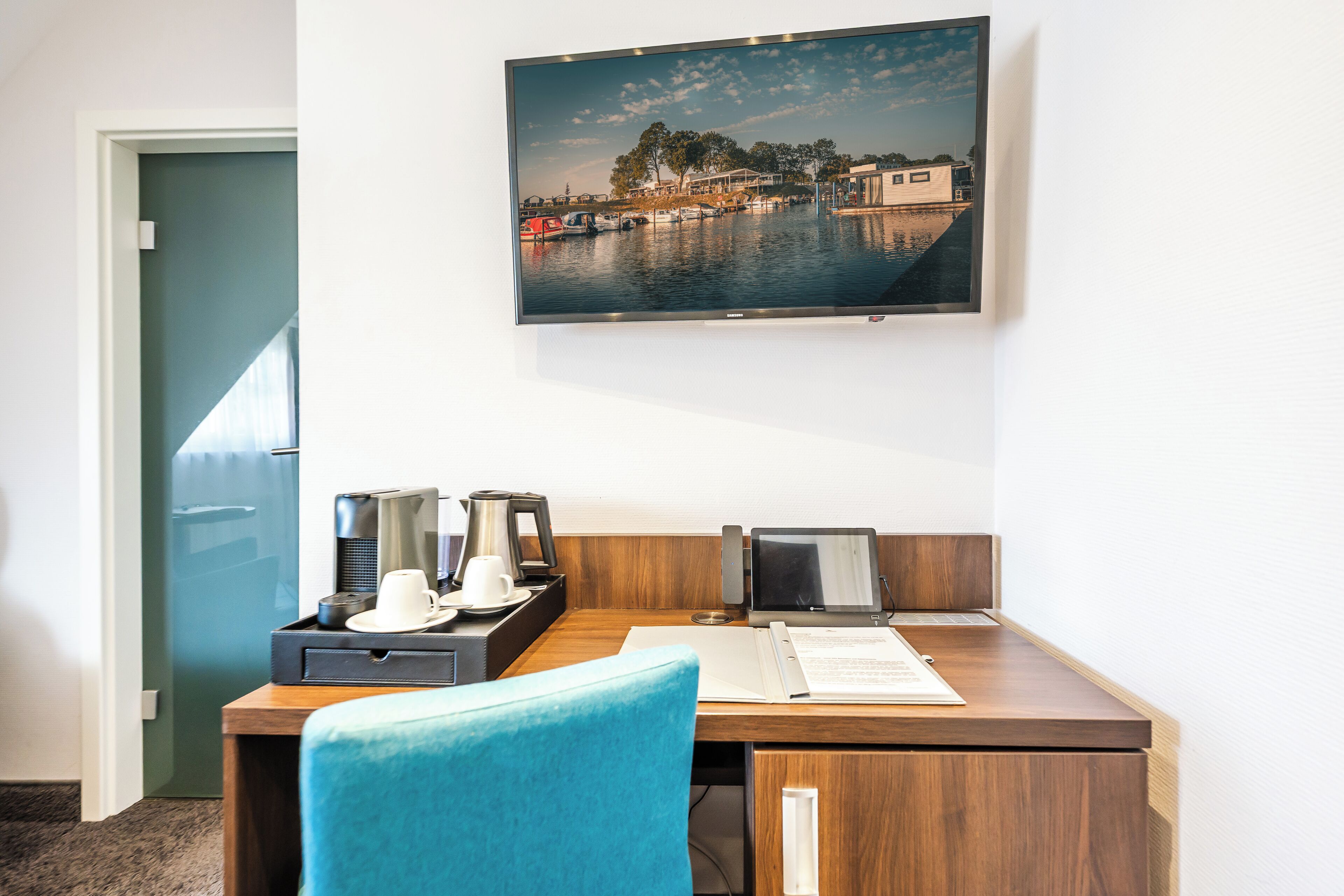 Een hotelkamer met een houten bureau, een blauwe stoel, een flatscreen televisie aan de muur en een deur die naar een andere kamer leidt.