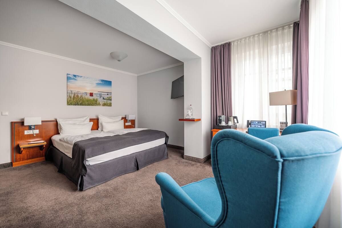 A hotel room with a bed, a desk, a chair, a TV, and a painting on the wall.