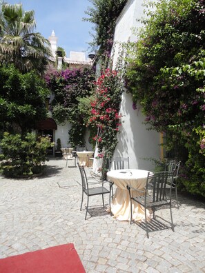 Un patio al aire libre con mesas y sillas, rodeado de abundante follaje y plantas con flores.