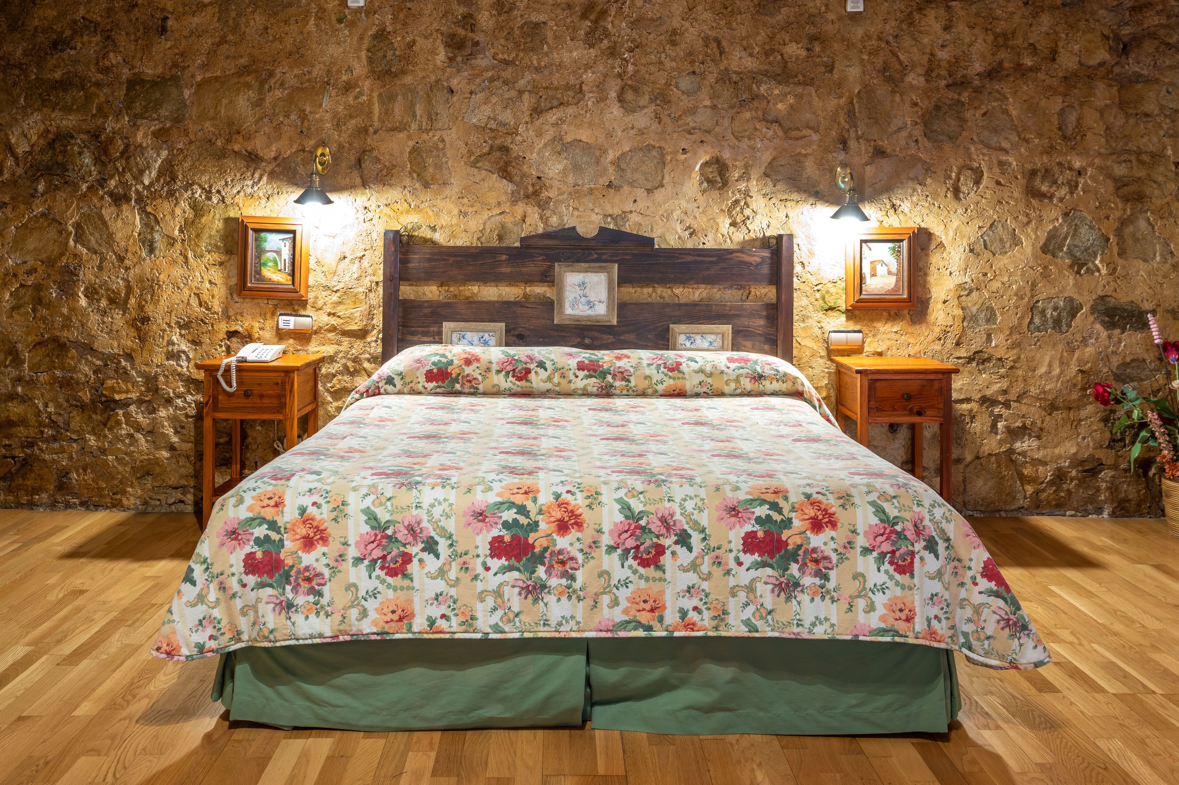 Un dormitorio con cabecera de madera, colcha con estampado floral, dos mesitas de noche con lámparas y una pared de piedra.