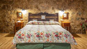 Un dormitorio con cabecera de madera, colcha con estampado floral, dos mesitas de noche con lámparas y una pared de piedra.