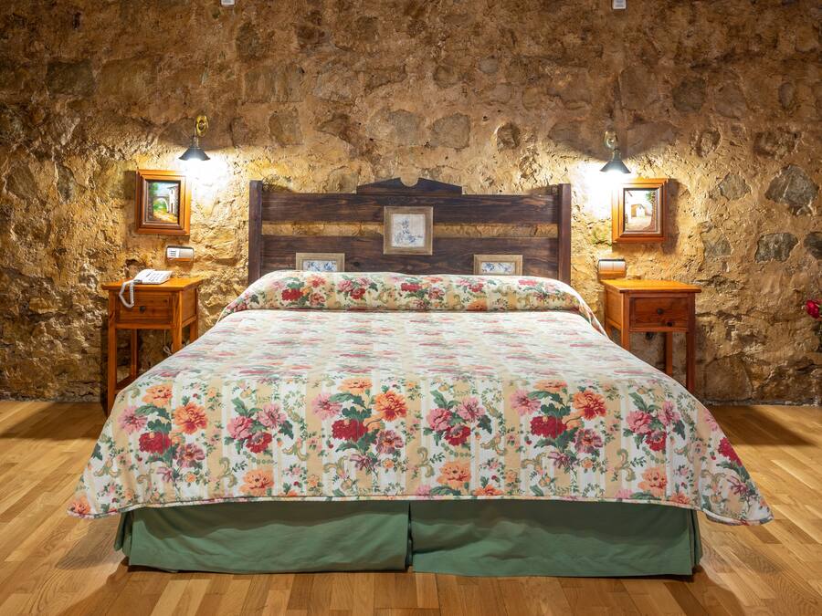 Un dormitorio con cabecera de madera, colcha con estampado floral, dos mesitas de noche con lámparas y una pared de piedra.
