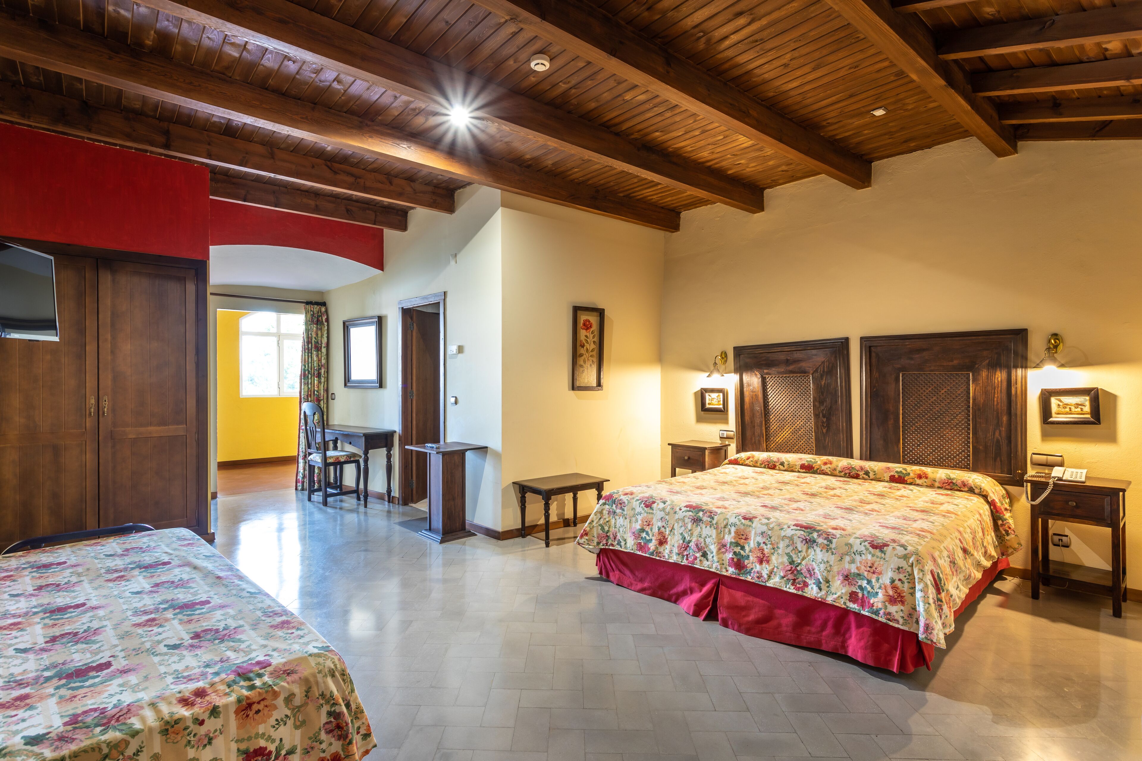 Habitación de hotel con techo de madera, cama grande con ropa de cama floral, escritorio con silla y una mesita.