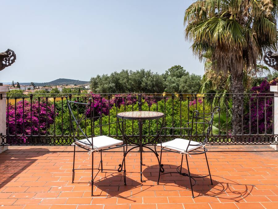 Una terraza con una mesa redonda y dos sillas, rodeada por una verja de hierro forjado, con vistas a un jardín lleno de plantas y colinas a lo lejos.