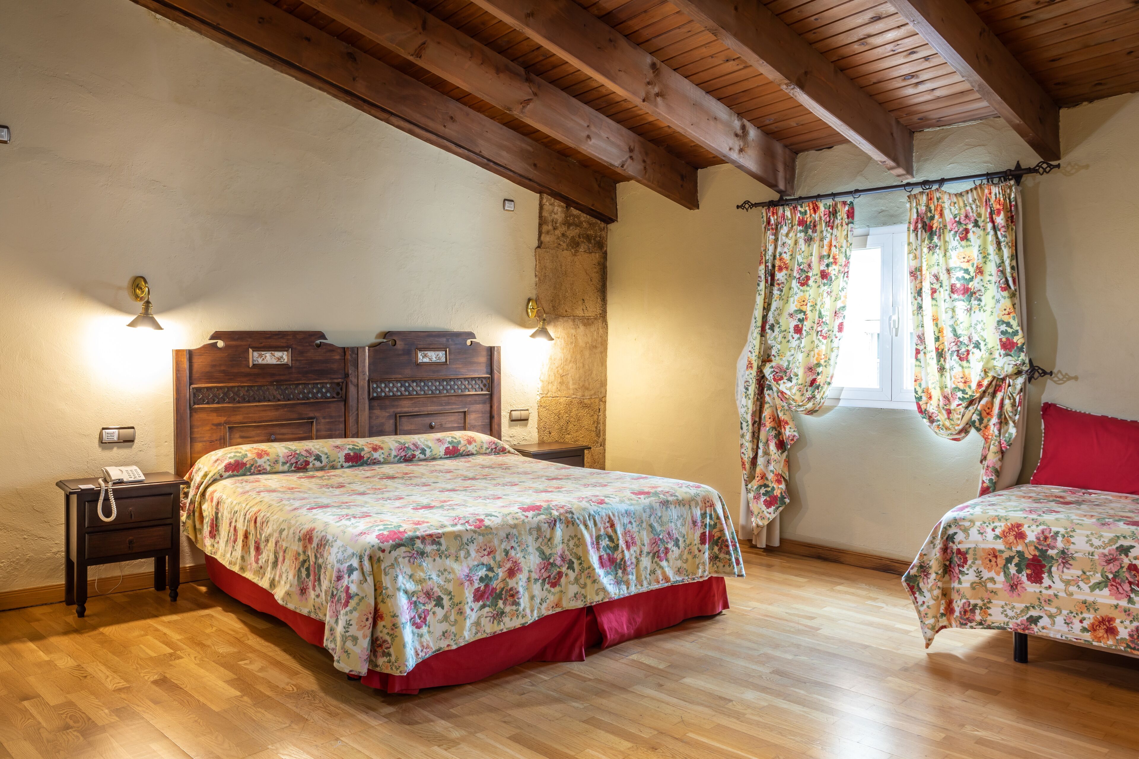 Une chambre avec deux lits, un plancher en bois, une tête de lit en bois, une table de chevet et une fenêtre agrémentée de rideaux à motifs floraux.