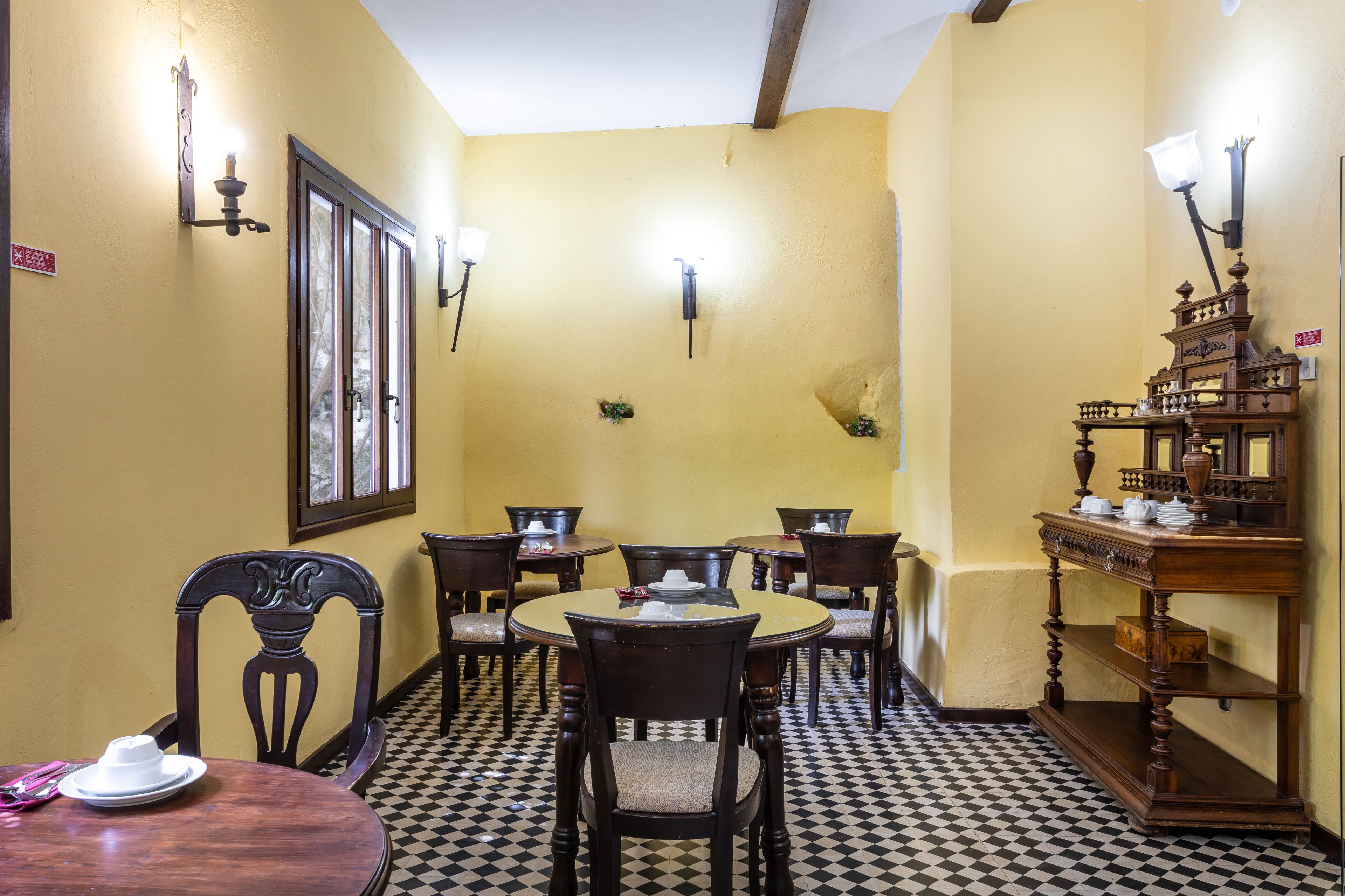 A dining area with a checkered floor, wooden chairs, a round table, and wall-mounted light fixtures.