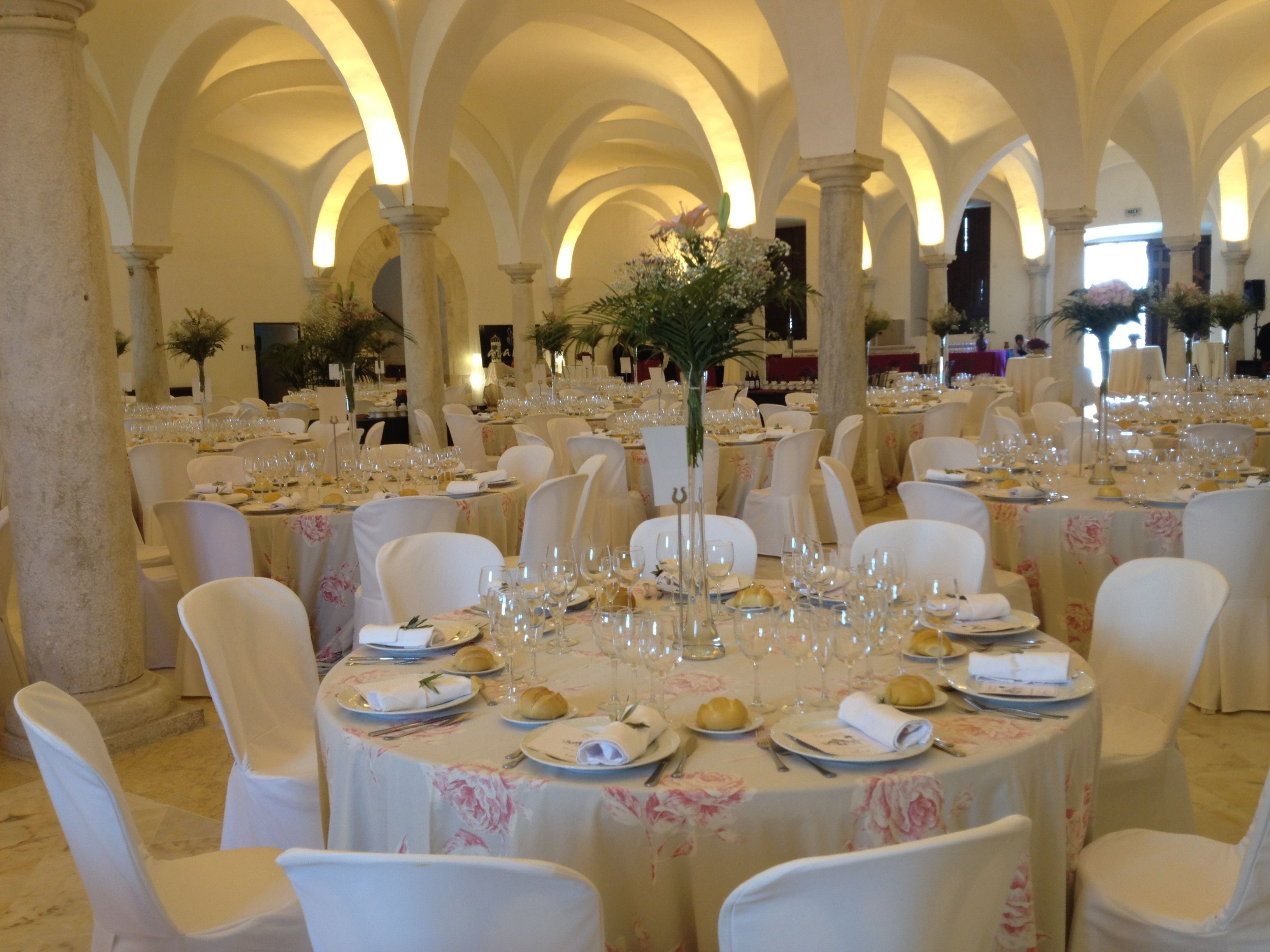 Un salón de banquetes con mesas redondas dispuestas para un evento formal, con sillas blancas y manteles florales.