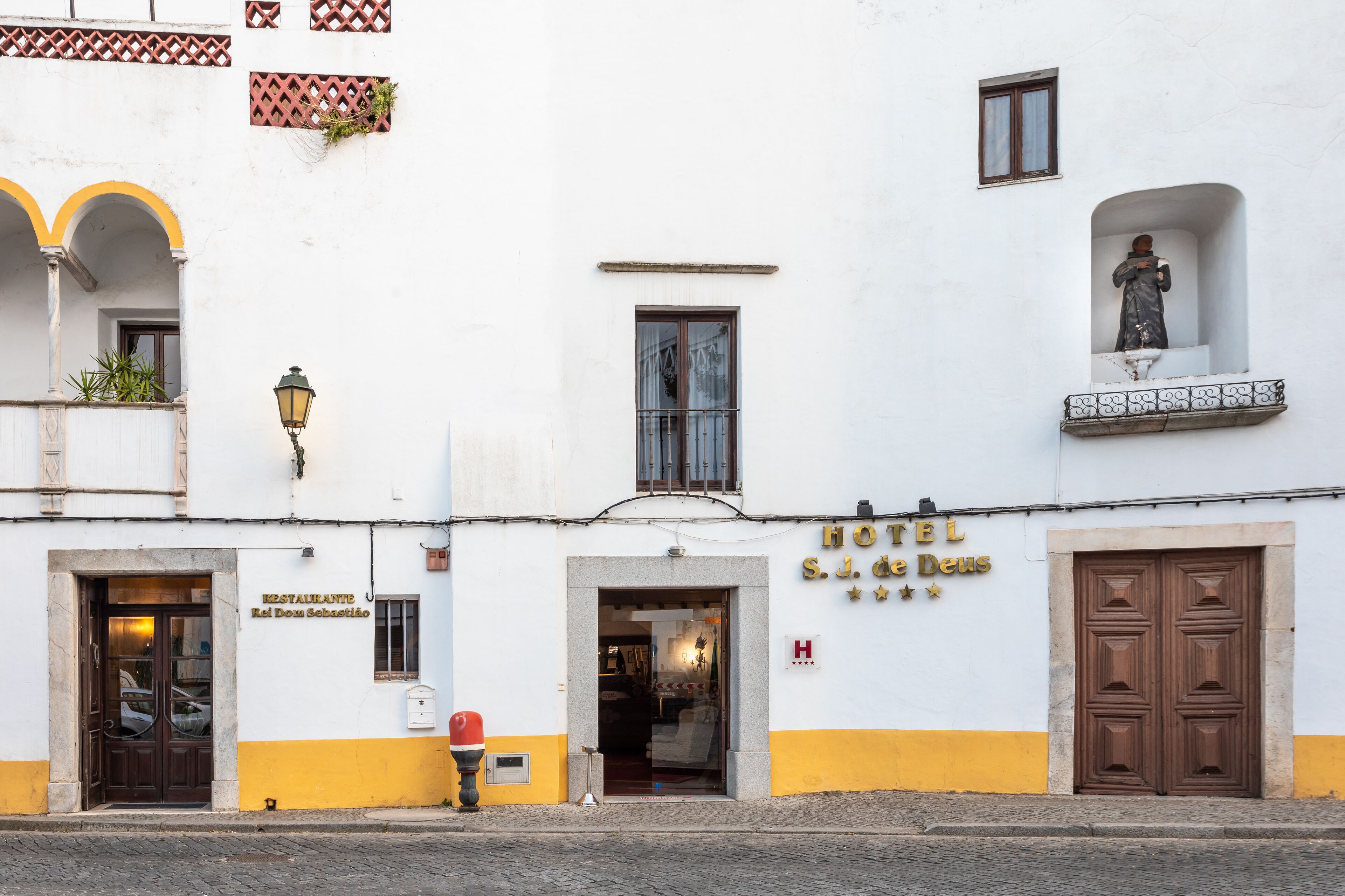 Un edificio blanco con base amarilla, un letrero que dice "HOTEL S.J. de Deus" y una estatua en la esquina superior derecha.